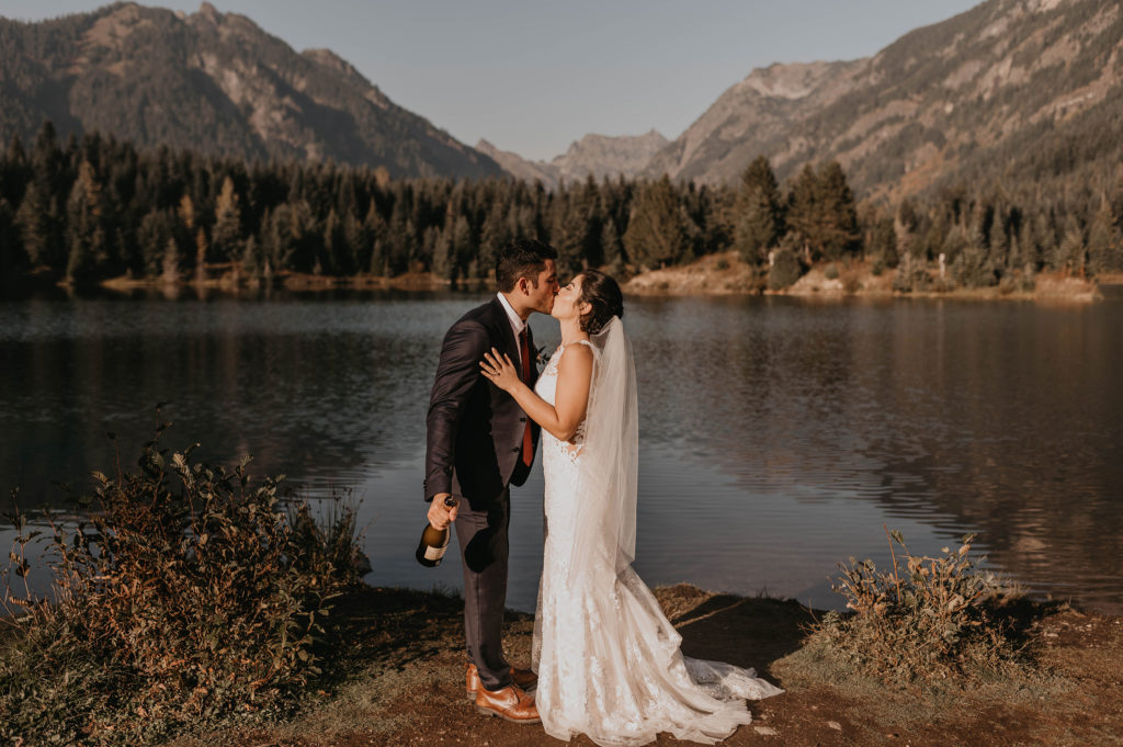 A newly married couple in wedding attire share a kiss after their Gold Creek Pond Elopement. Featuring an alpine lake in front of mountains.
