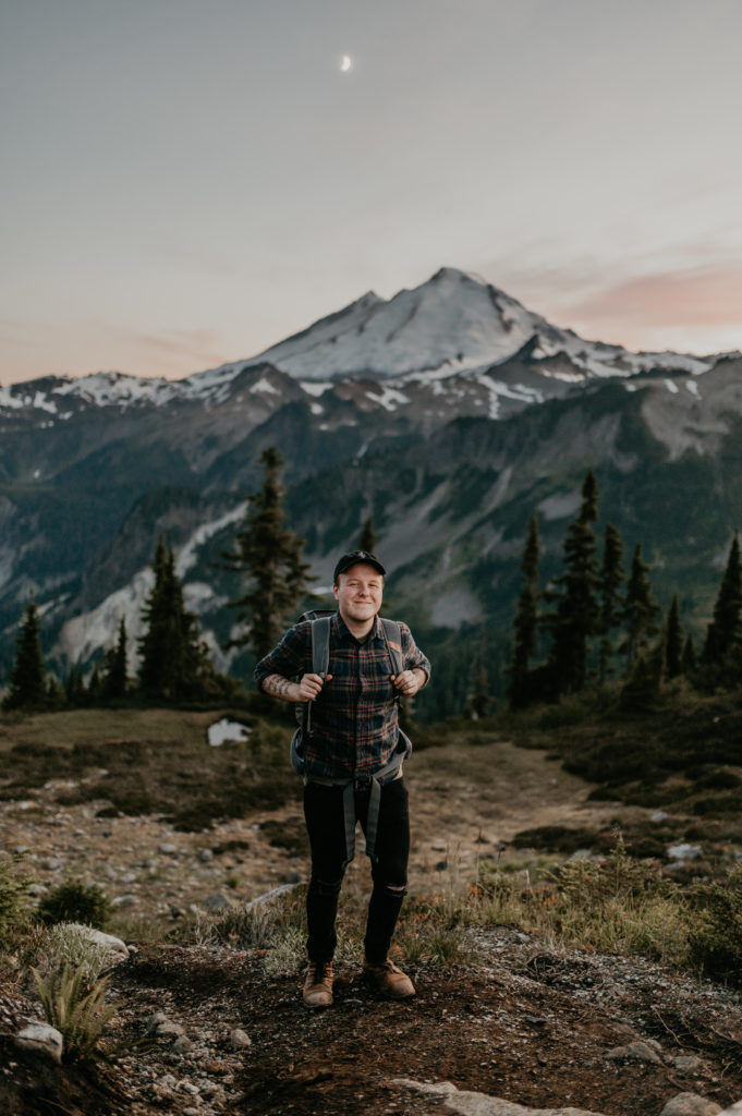An Olympic National Park photographer poses on a trail in front of a mountain.
