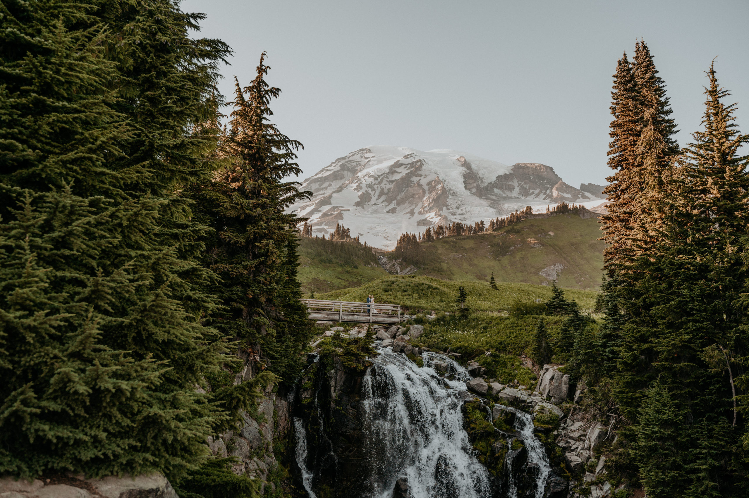 Couple celebrates their Mount Rainier Elopement while standing on a bridge above a waterfall in front of Mount Rainier.