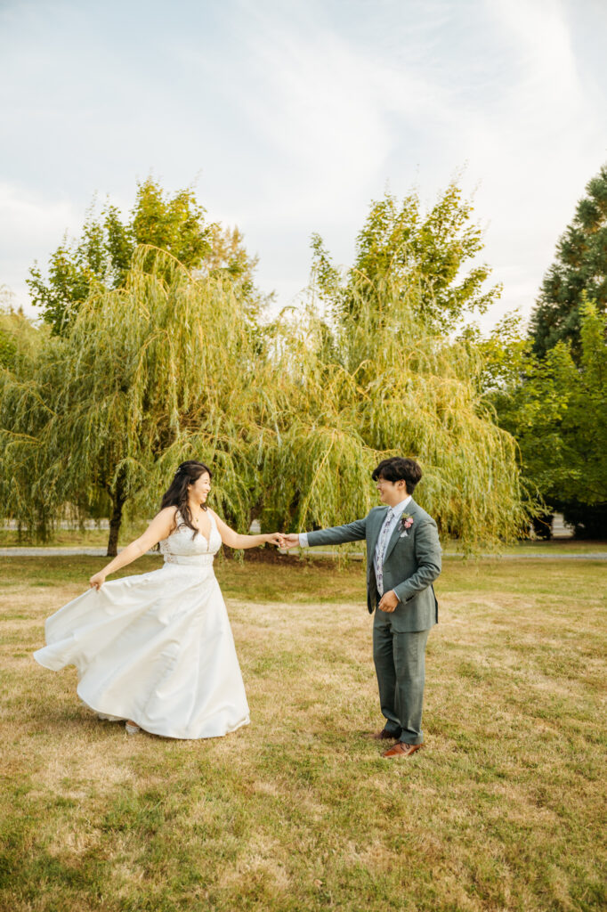 Bride and groom dance in a field in front of a willow tree while taking golden hour photos at Twin Willow Gardens